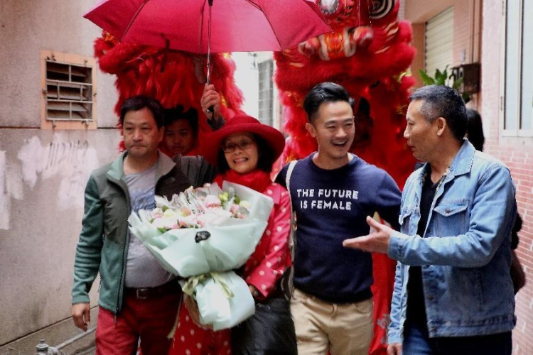 Jenny and Ben are led by villagers down a local road with a bouquet of flowers and lion dancers escorting her from behind
