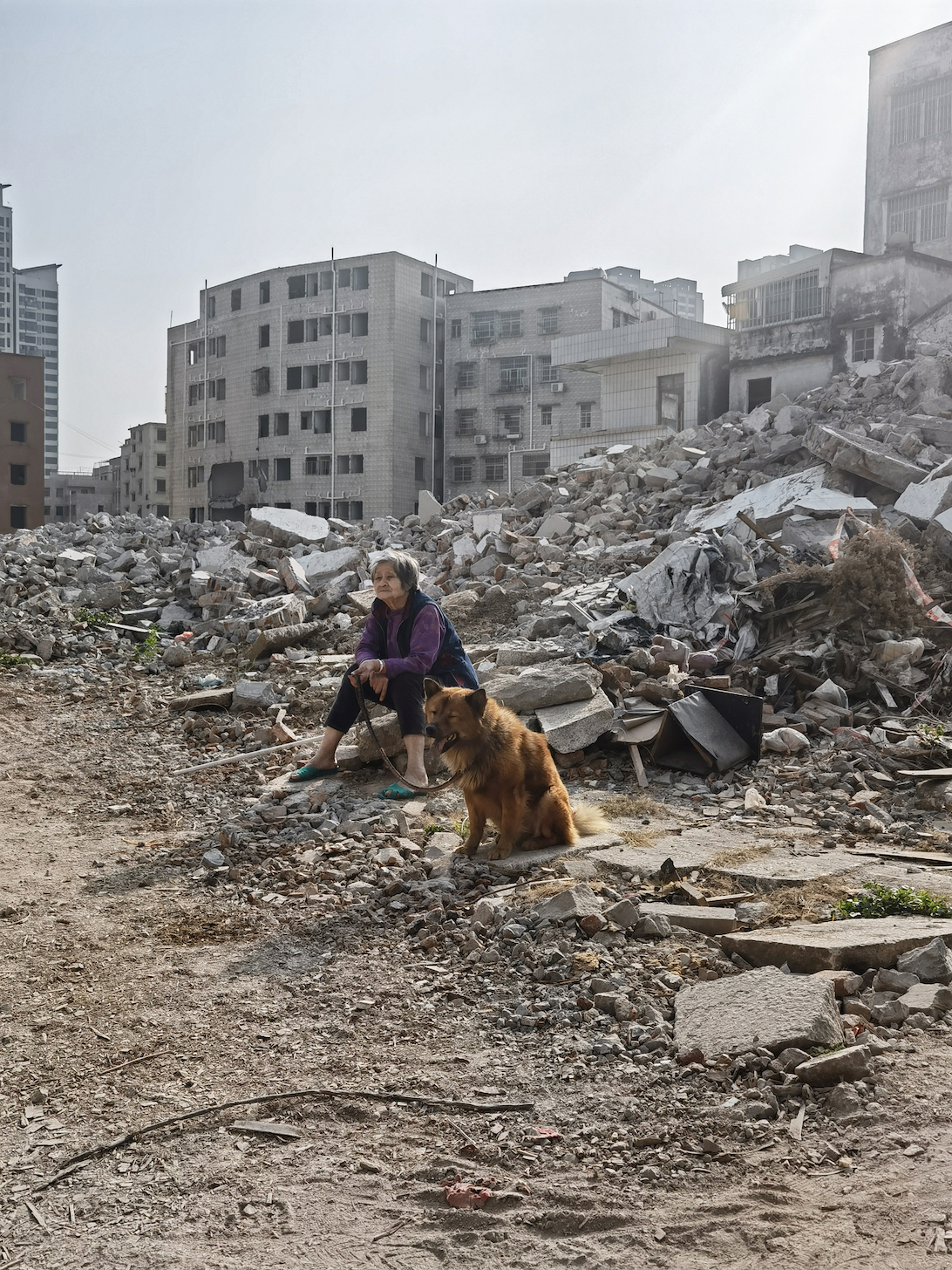 An elderly Chinese woman sits amongst rubble with her dog, looking pensive