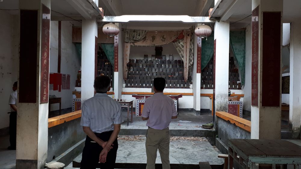 Two men standing in a dimly lit ancestral hall, their backs to the camera, with the ancestral tablets in the background.
